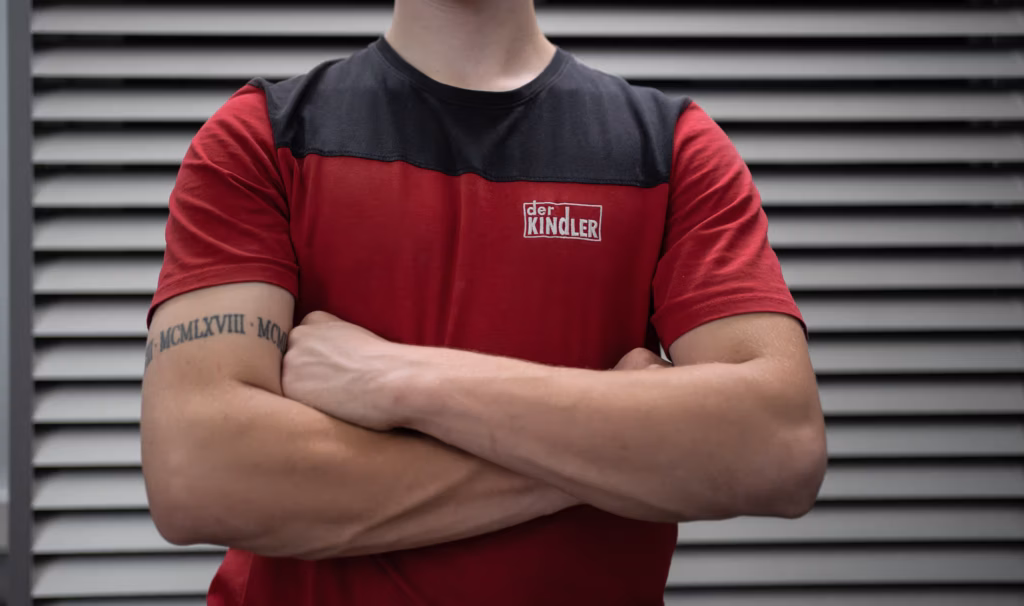 Nahaufnahme eines Mitarbeiters mit verschränkten Armen im Firmen-Shirt von ‚Der Kindler‘ – Corporate Fotografie im Handwerk